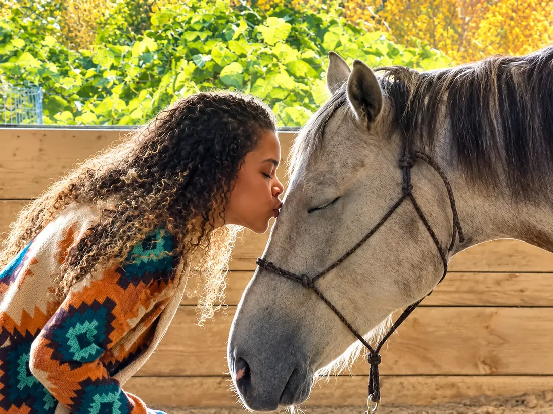Alyson with her eyes closed kissing a calm gray horse on the forehead in a sunlit arena, with green and golden trees behind