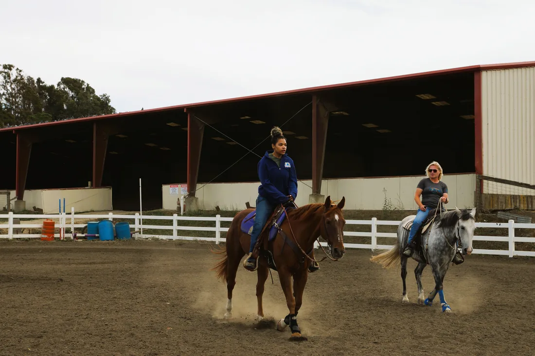 Alyson riding a chestnut horse through a dusty arena while another rider on a gray horse follows behind, with a red covered arena in the background