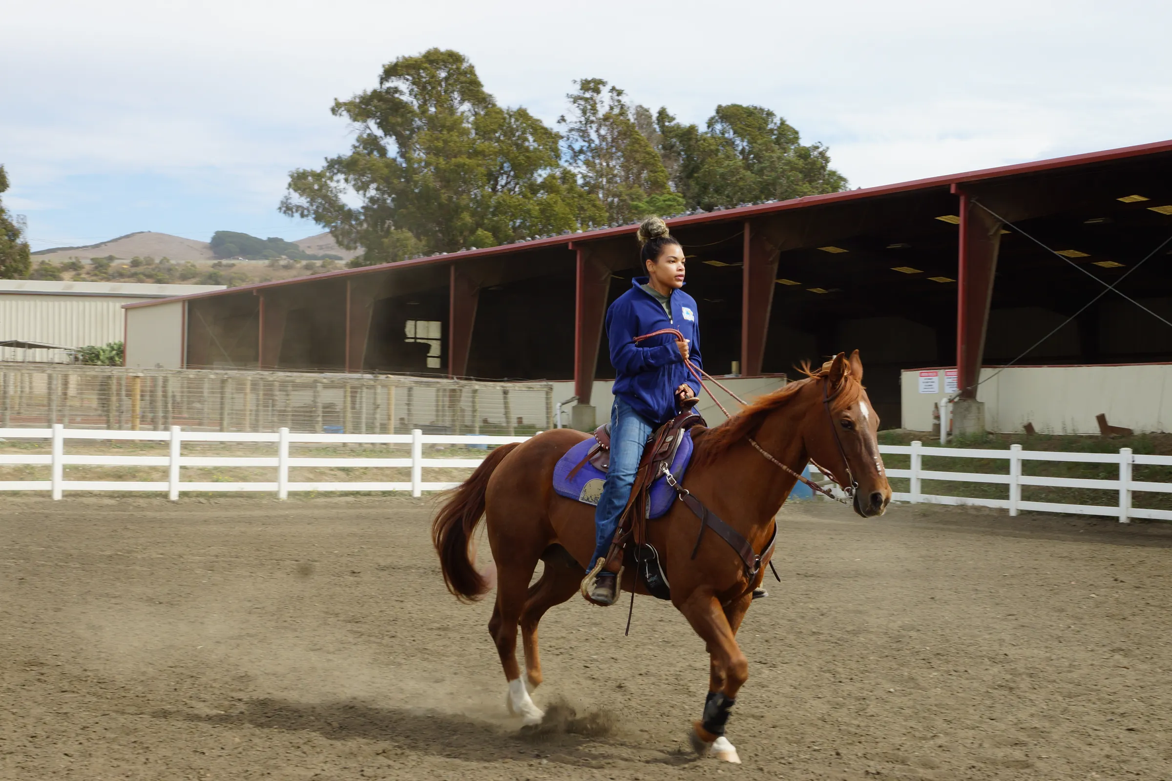 Alyson in a blue jacket riding a chestnut horse at a lope across a dirt arena, with a white fence and a red covered arena behind