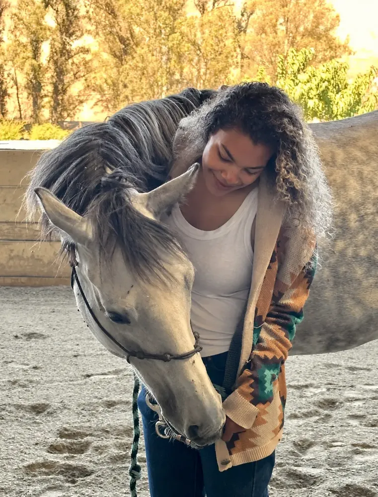 Alyson leaning over a gray horse as it rests its head against her in a sunlit arena