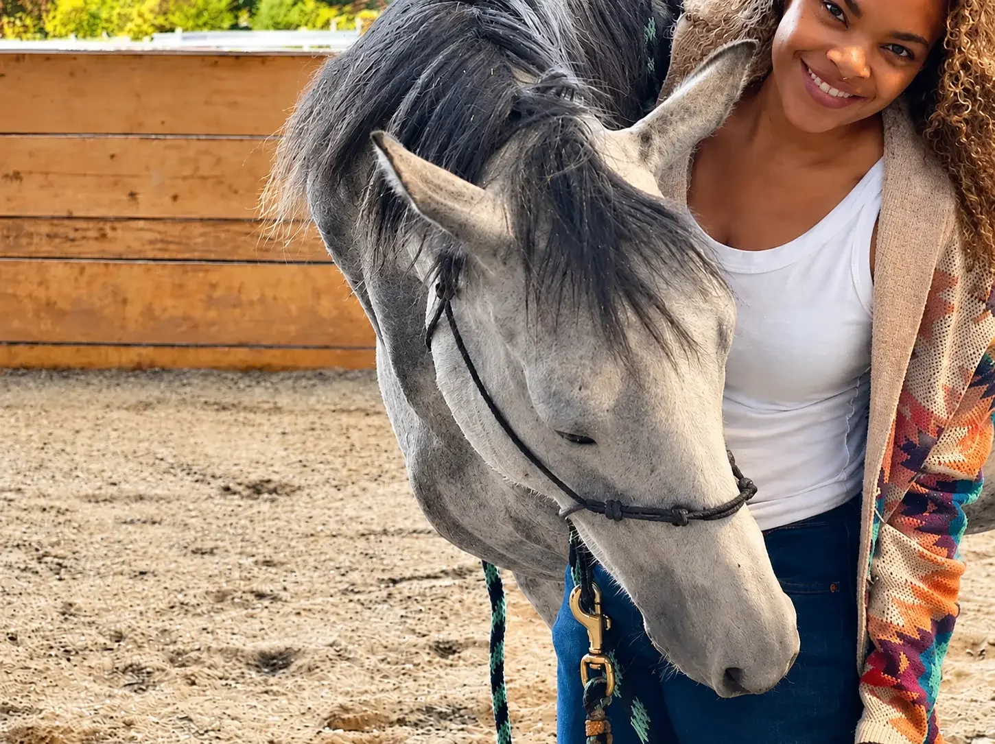 Alyson smiling beside a relaxed gray horse as it lowers its head with eyes closed in a sandy arena, with a wooden fence behind